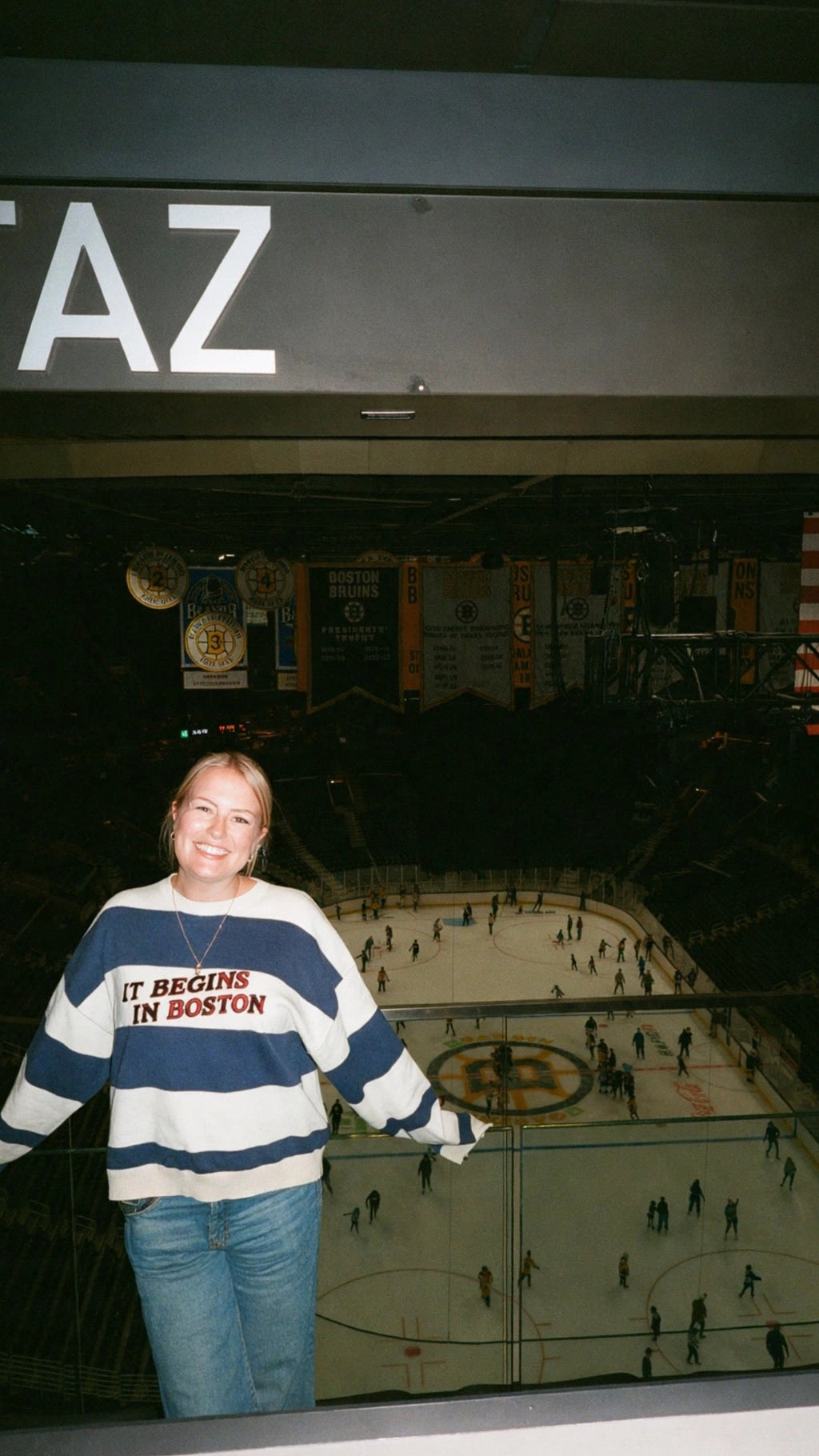 A woman wearing a navy and white striped 'It Begins in Boston' sweater stands in an indoor arena overlooking an ice hockey rink.