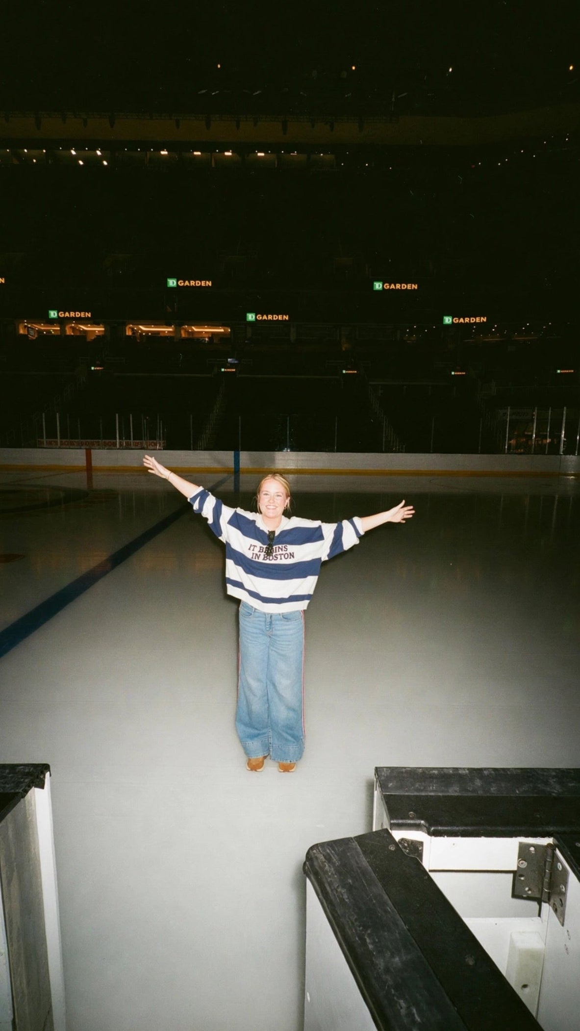 A person stands on an ice rink with arms outstretched, wearing a navy and white "It Begins In Boston" striped long-sleeve top and light blue jeans in a dark indoor arena.