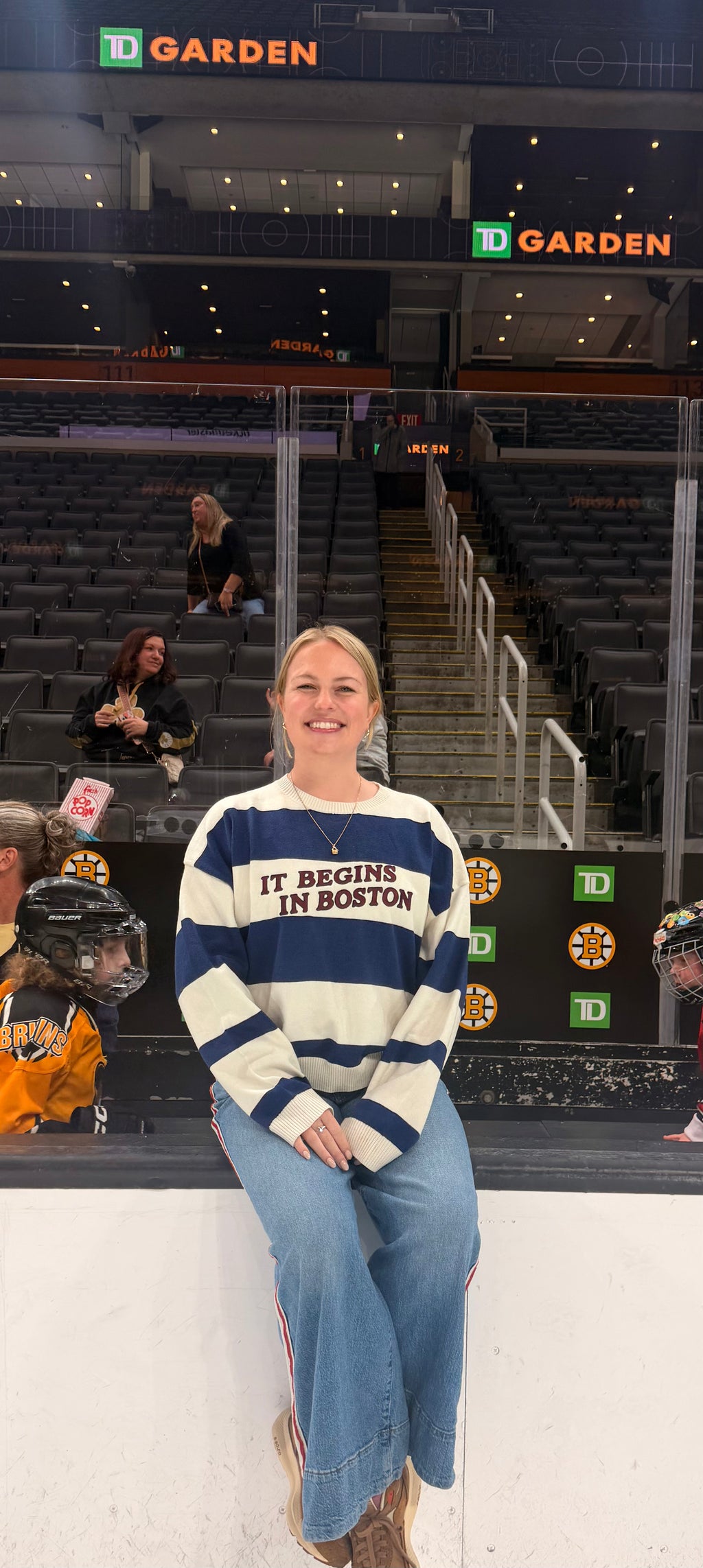 A woman sitting on the ice at TD Garden wearing a navy and cream striped sweater with 'IT BEGINS IN BOSTON' across the chest, paired with blue jeans.