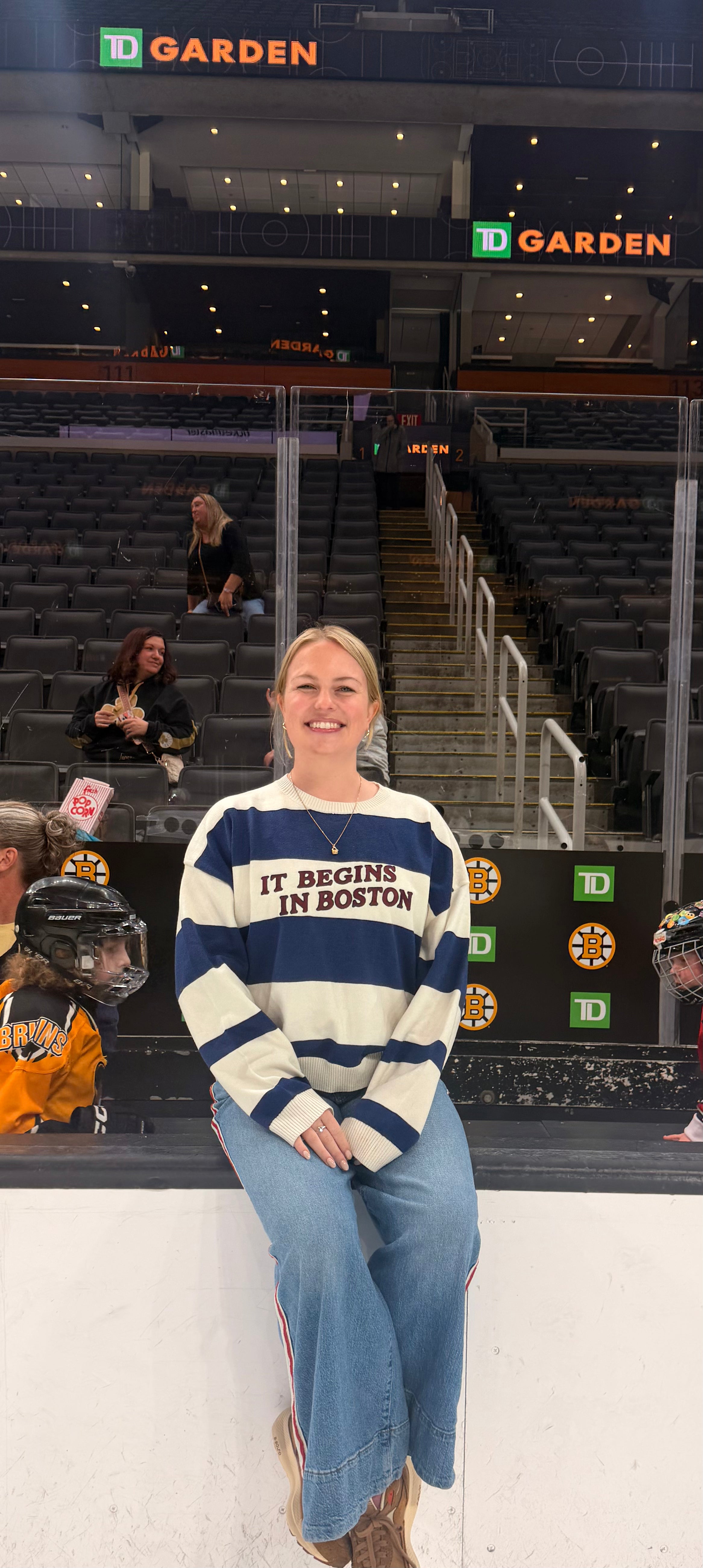 A woman sitting on the ice at TD Garden wearing a navy and cream striped sweater with 'IT BEGINS IN BOSTON' across the chest, paired with blue jeans.